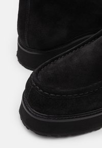 Stivaletti neri in suede con punta arrotondata, cuciture a contrasto e una sola in gomma spessa. Vista ravvicinata che mette in evidenza la texture e i dettagli del design.