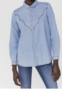Light blue denim shirt with a button-up front, pointed collar, and decorative seam accents. Long sleeves with buttoned cuffs.