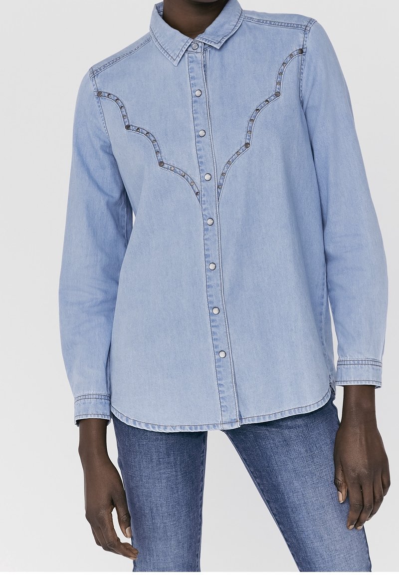 Light blue denim shirt with a button-up front, pointed collar, and decorative seam accents. Long sleeves with buttoned cuffs.