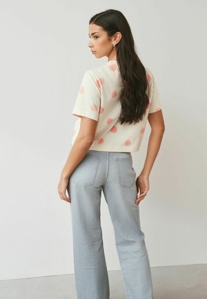 Hosszú, sötét hajú nő, fehér, rövid felsőt visel rózsaszín pöttyökkel és világoskék, magas derekú farmernadrágot, egy sima falnak támaszkodva áll.