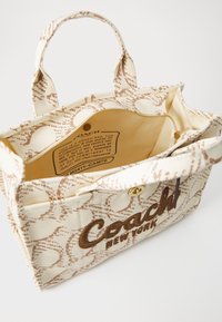 Canvas handtas in crème, met een bruine logo, zigzagpatroon en twee korte handvatten. Het interieur heeft een ritsvak en is gevoerd.