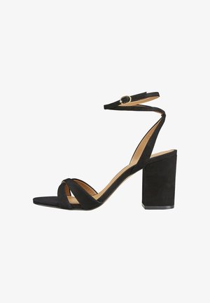 Sandale à talon haut en daim noir avec talon bloc, bride à la cheville avec boucle dorée, et brides croisées sur les orteils.