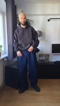 Moška siva kapuca z sprednjim žepom, v kombinaciji z ohlapnimi temno modrimi jeans hlačami in črnimi čevlji. Preprost, sodoben dizajn z minimalnimi poudarki.
