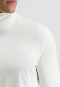 Close-up van een persoon die een witte coltrui draagt met een klein "pierre cardin" logo op de borst.