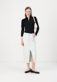 Haut zippé noir à manches longues, jupe midi en denim bleu clair avec une fente sur le devant, et chaussures plates noires. Tient un petit sac à main rose.