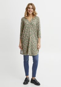 Floral Tunika-Kleid in Grün, mit V-Ausschnitt, Dreiviertelärmeln und lockerer Passform. Der Rock hat seitliche Schlitze und wird mit blauen Jeans und schwarzen Clogs kombiniert.
