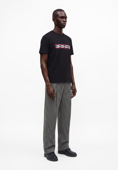 Homme portant un t-shirt noir avec un texte graphique, pantalon large gris à fines rayures et chaussures noires, debout devant un fond blanc uni.