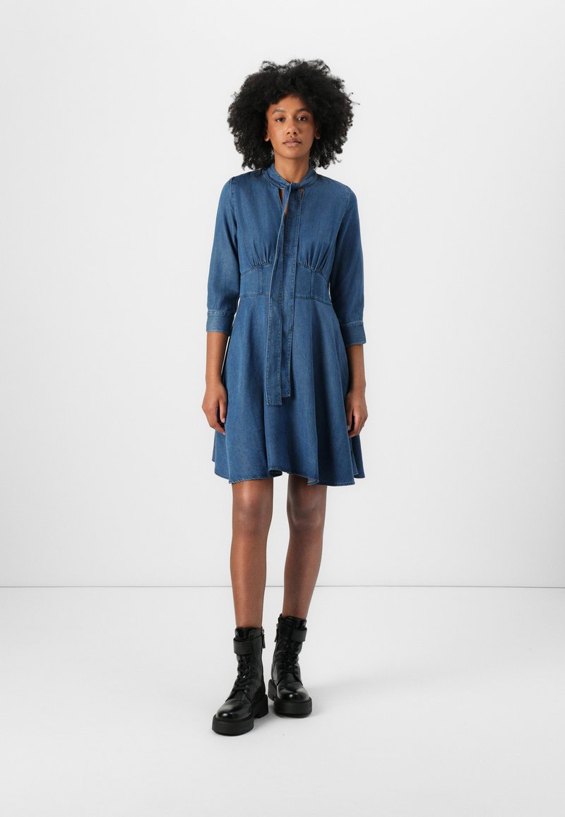 Denim dress with a collar, pleated waist, and three-quarter sleeves. Lined pockets and a flowing hem. Worn with black lace-up boots.