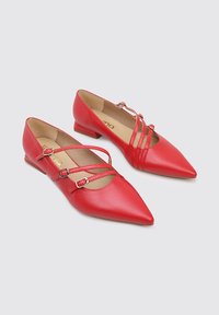 Bailarinas rojas de imitación cuero con punta afilada, que presentan dos adornos de tira ajustables y un tacón bajo. Textura suave y diseño minimalista.