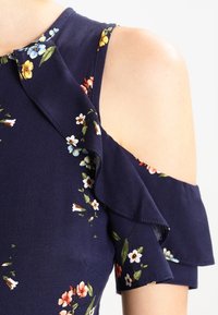 Granatowa sukienka w kwiaty z odkrytymi ramionami, falbanującymi rękawami oraz teksturowanym materiałem w wielokolorowe wzory kwiatowe.