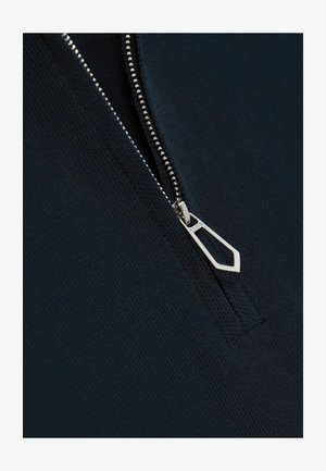 Giaccone navy con zipper, realizzato in un tessuto strutturato, con una zip argentata dotata di un design geometrico. Vista ravvicinata del dettaglio della zip.