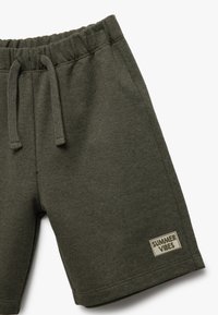 Grüne Baumwollshorts mit elastischem Bund und Kordelzug. Merkmale ein beiges Etikett "SUMMER VIBES" auf der unteren linken Seite.