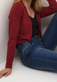 Rode aansluitende cardigan over een zwart shirt, gecombineerd met blauwe spijkerbroek. De cardigan heeft een zachte textuur, terwijl de jeans subtiele stiksels bevatten.
