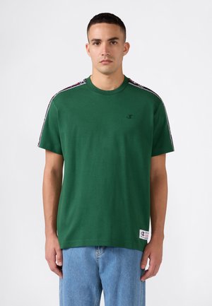 Man draagt een groen kortemouw T-shirt met logoband op de schouders en lichtblauwe jeans, staand tegen een effen achtergrond.
