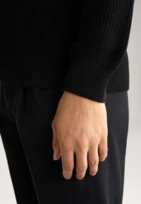 Schwarzer Strickpullover mit geripptem Muster und engen Bündchen, kombiniert mit dunklen Hosen; eine Hand ist sichtbar und ruht gegen die Kleidung.