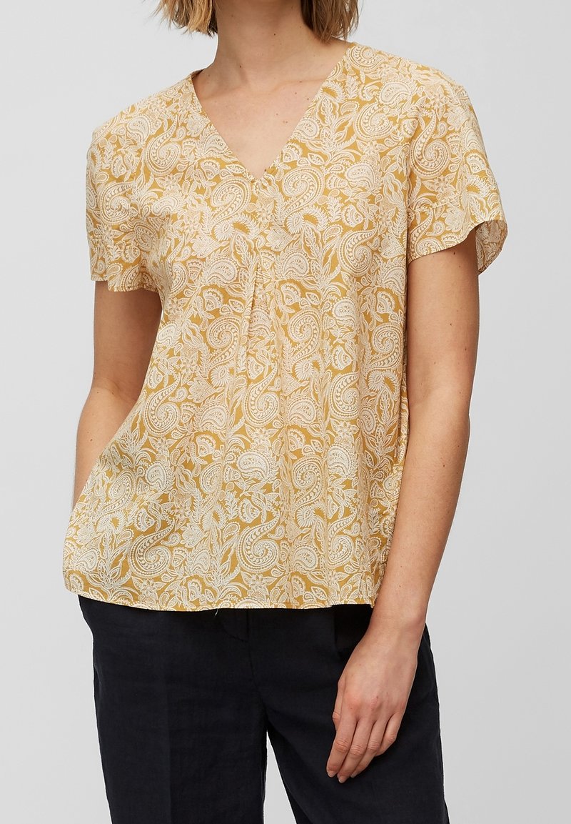 Femme portant un chemisier jaune à manches courtes avec motif cachemire blanc et col en V, associé à un pantalon noir, debout devant un fond uni.