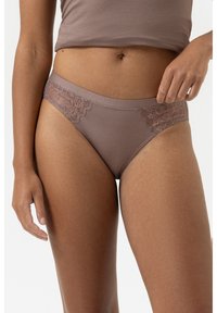 Frau in mauve-farbenem Unterwäsche-Bikini mit Spitzenpaneel und passendem Crop-Top, stehend vor einem schlichten weißen Hintergrund.