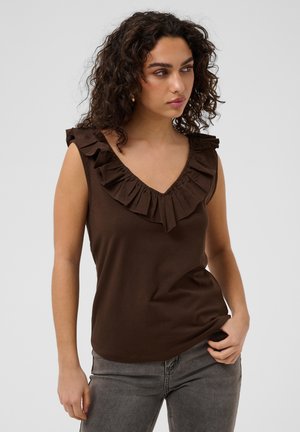 Femme aux cheveux bouclés portant un haut marron sans manches avec un col en V volanté et un jean gris, debout devant un fond clair uni.