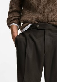 Maglione marrone lavorato a maglia con maniche arrotolate abbinato a pantaloni scuri, sartoriali, con chiusura a bottone e tasche laterali.