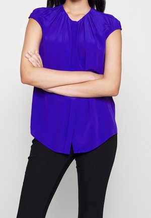 Frau mit langem dunklem Haar, die eine lila Bluse mit kurzen Ärmeln und schwarze Hose trägt, steht mit verschränkten Armen vor einem schlichten Hintergrund.