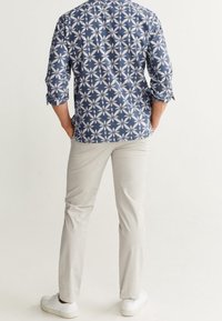 Camicia a maniche lunghe da uomo, con motivo floreale blu e bianco, in tessuto di cotone. Abbinata a pantaloni chiari e sneakers bianche.