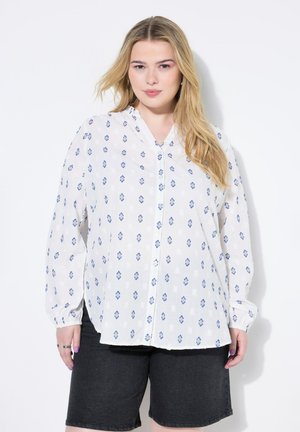 Weiße Bluse mit blauer geometrischer Musterung, langen Ärmeln und V-Ausschnitt, kombiniert mit dunklen Jeansshorts. Leichtes, transparentes Material.
