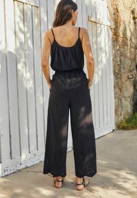 Schwarzer weit geschnittener Jumpsuit mit einem Schlüssellochrücken, bestickten Details und Lochmuster-Saum. Hergestellt aus leichtem Stoff, kombiniert mit braunen Sandalen.