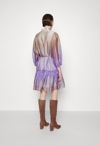 Robe en tulle dégradé passant du mauve au violet, avec des manches bouffantes, une taille cintrée et un ourlet volanté. Associée à des bottes hautes en daim marron.