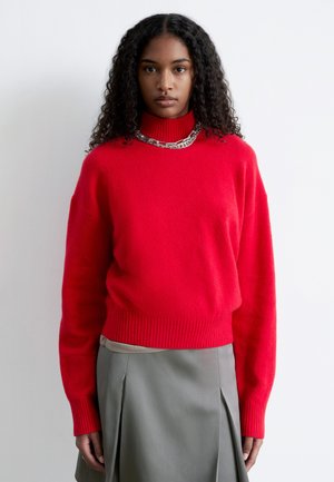 Rød uldtrøje med ribbet halsudskæring og manchetter, kombineret med et gråt plisseret skørt. Stoffet virker blødt med en glat tekstur.