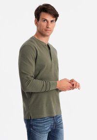 Junger Mann in olivgrünem Langarm-Henley-Shirt und blauer Jeans, der vor einem weißen Hintergrund steht und in die Kamera schaut.