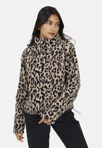 Donna che indossa un pullover in pile leopardato beige e nero con mezza zip, in piedi con una mano vicino alla vita, mentre guarda di lato.