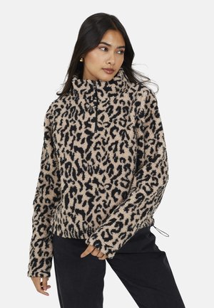 Vrouw in een beige en zwarte panterprint fleece pullover met een halve rits, staand met één hand bij de taille, kijkend naar de zijkant.