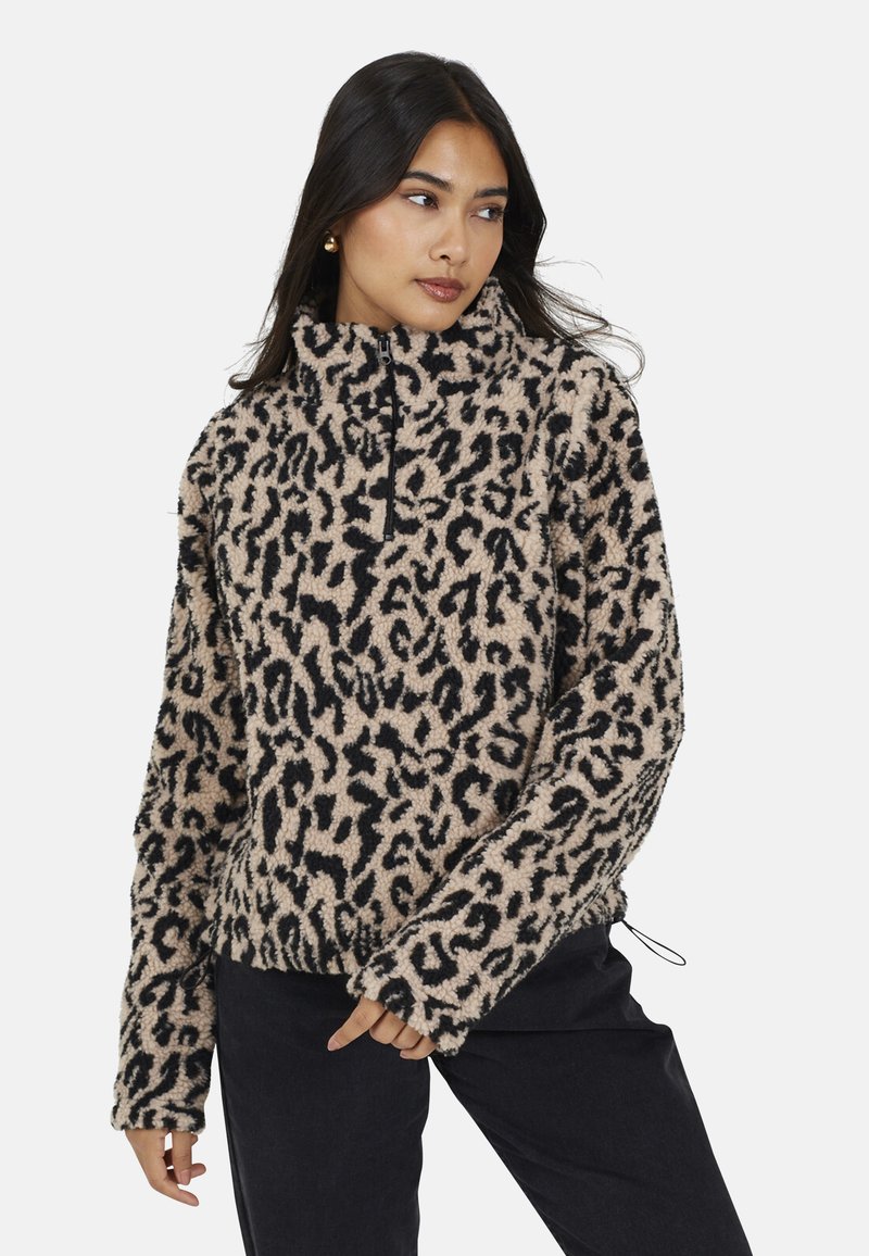 Donna che indossa un pullover in pile leopardato beige e nero con mezza zip, in piedi con una mano vicino alla vita, mentre guarda di lato.