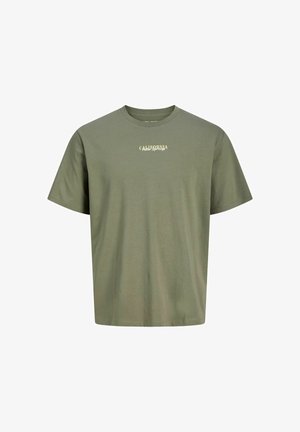 Tiener met krullend haar, gekleed in een olijfgroen "California" T-shirt, losse blauwe spijkerbroek en bruine schoenen, staand tegen een witte achtergrond.