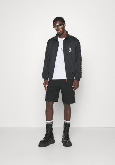 Μαύρο μπουφάν φόρμας με λογότυπο, λευκό μπλουζάκι, μαύρες βερμούδες και μαύρες μπότες πλατφόρμας. Διακοσμητικά στοιχεία περιλαμβάνουν χρυσό σχέδιο στο πλάι και ριγέ κάλτσες.