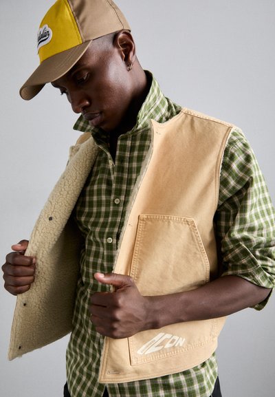 Tan shearling-lined vest, green checkered shirt, and beige cap with a yellow brim. Vest features a pocket and subtle logo detail.