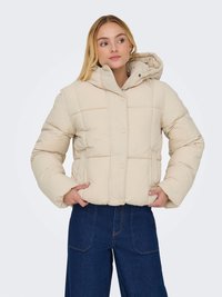 Casaco bege almofadado com um colarinho alto e botões de pressão. Apresenta um design acolchoado e um capuz, usado sobre calças de denim azul-escuro.