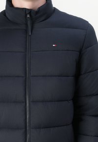 Marineblå pufferjakke med horisontale quiltede detaljer, høy krage, glidelås foran og liten logo på brystet. Glatt, lett stoff.