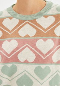 Tröja med mintgrön krage som har rader av vita hjärtan på en flätad tyg med tan, rosa och mintgrön zick-zack mönster.