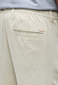 Közeli kép világos bézs színű, texturált nadrágról, melynek elasztikus derékrésze és hátsó hasítékos zsebe van, kis Jack & Jones logó címkével.