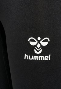 Leggings neri attillati realizzati in tessuto liscio, dotati di un logo "hummel" bianco e un design grafico sopra il logo.
