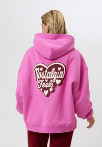 Felpa rosa con una grande grafica a forma di cuore marrone che presenta il testo "Nostalgia Feels", circondata da piccoli cuori bianchi. Vestibilità morbida e rilassata.