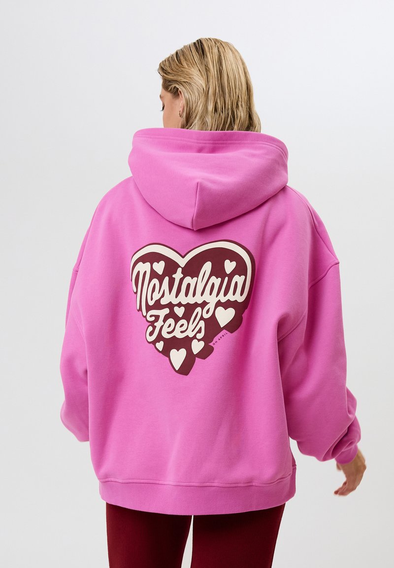 Felpa rosa con una grande grafica a forma di cuore marrone che presenta il testo "Nostalgia Feels", circondata da piccoli cuori bianchi. Vestibilità morbida e rilassata.
