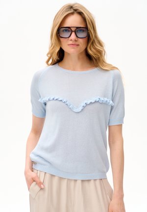 Kobieta o długich blond włosach, nosząca duże okulary przeciwsłoneczne w oprawkach w kolorze żółwia, jasnoniebieski krótki sweter z falbankami oraz beżowe spodnie.