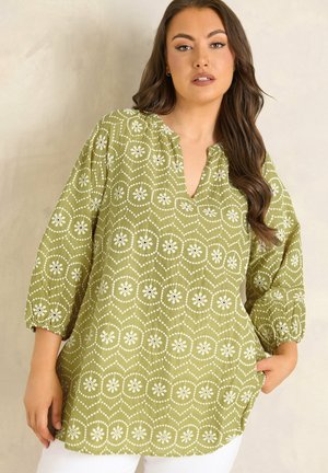 Vrouw met lang bruin haar die een groene tuniek draagt met witte bloemenpatronen en driekwartmouwen, poserend met één hand in de zak tegen een neutrale achtergrond.