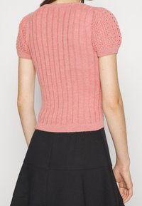Suéter de punto rosa con mangas cortas abullonadas, que presenta una textura acanalada y un detalle de calado en las mangas, combinado con una falda negra.