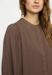 Bluză lungă, cu mâneci lungi, de culoare marron, cu un guler înalt, având accente plisate la umeri, plachetă cu butoni și material texturat neted.