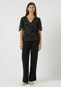 Zwarte blouse van bloemenkant met een V-hals en pofmouwen, gecombineerd met zwarte broek met wijde pijpen en lichtgekleurde schoenen.