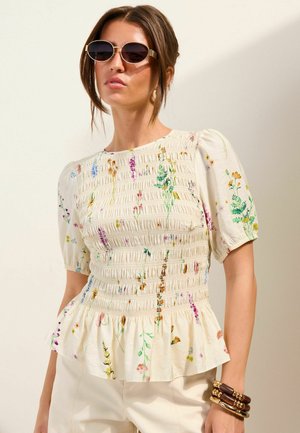 Naine, kes kannab ümmarguseid päikeseprille, kreemjat lillelist kroogitud pluusi lõhnavate varrukatega ja kihilisi puidust käevõrusid randmel.