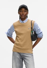 Kobieta z warkoczami, ubrana w beżowy sweter na koszulę w niebieską kratę, szare dżinsy, czapkę w kolorze czarnym i nosząca czarną torbę na ramię.